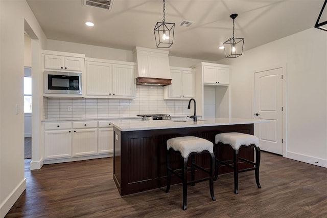 kitchen with dark wood-type flooring, an island with sink, hanging light fixtures, custom range hood, and built in microwave