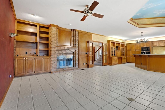 unfurnished living room with light tile patterned flooring, ceiling fan with notable chandelier, ornamental molding, a brick fireplace, and built in shelves