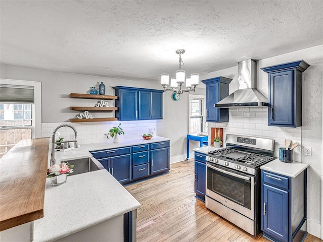 kitchen with wall chimney range hood, sink, blue cabinetry, stainless steel gas range oven, and decorative backsplash