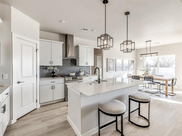 kitchen with white cabinetry, wall chimney exhaust hood, stainless steel range with gas cooktop, and a center island with sink