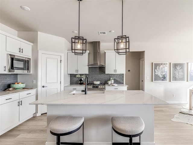 kitchen featuring pendant lighting, wall chimney range hood, white cabinetry, a center island with sink, and built in microwave