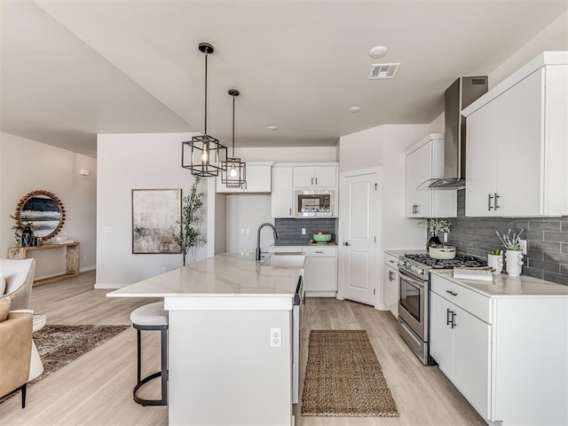 kitchen with wall chimney range hood, appliances with stainless steel finishes, a kitchen island with sink, light hardwood / wood-style floors, and white cabinets