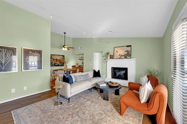 living area featuring visible vents, wood finished floors, baseboards, a brick fireplace, and ceiling fan