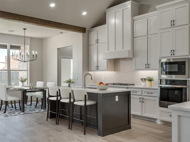 kitchen featuring appliances with stainless steel finishes, beam ceiling, tasteful backsplash, white cabinets, and a center island with sink