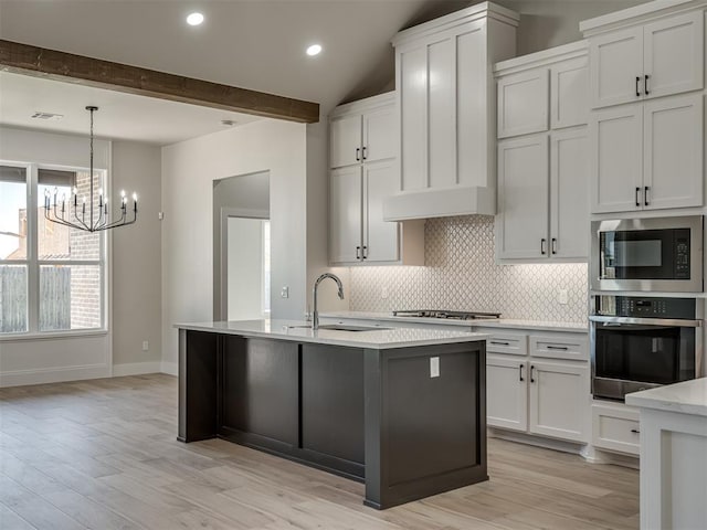 kitchen featuring appliances with stainless steel finishes, white cabinetry, backsplash, a center island with sink, and beam ceiling