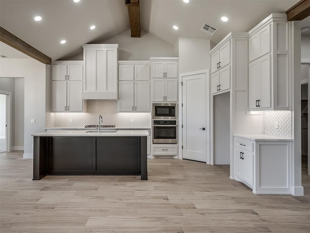 kitchen with white cabinetry, a kitchen island with sink, beam ceiling, black microwave, and stainless steel oven