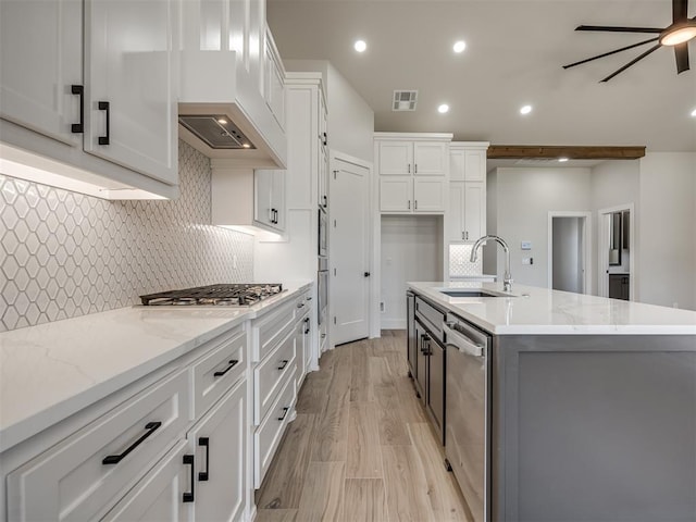 kitchen featuring sink, white cabinetry, stainless steel appliances, light stone counters, and a center island with sink