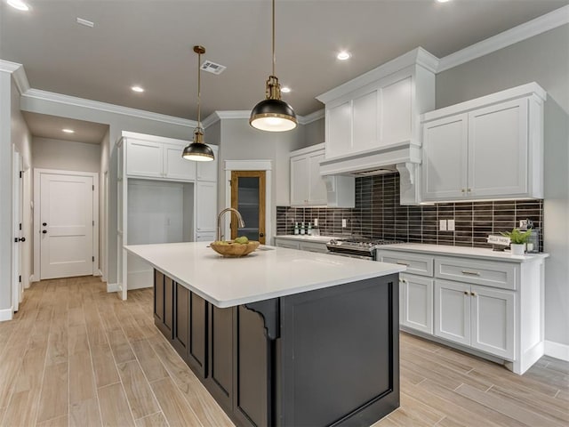 kitchen featuring white cabinetry, backsplash, hanging light fixtures, a kitchen island with sink, and crown molding