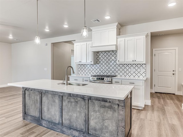 kitchen featuring sink, hanging light fixtures, an island with sink, stainless steel stove, and white cabinets