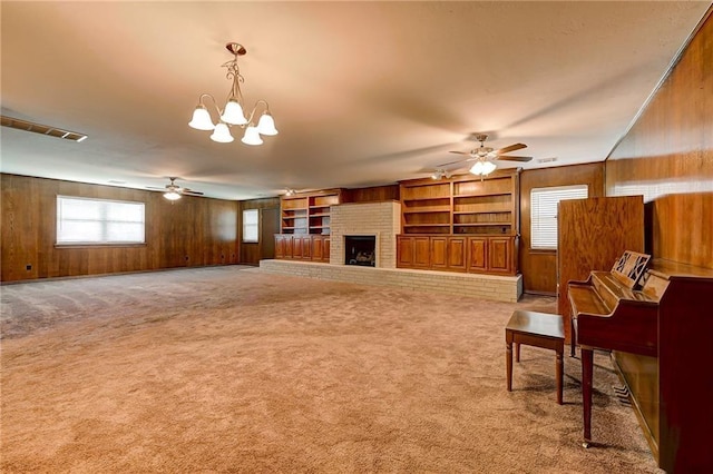 carpeted living room featuring ceiling fan with notable chandelier, built in features, wood walls, and a fireplace