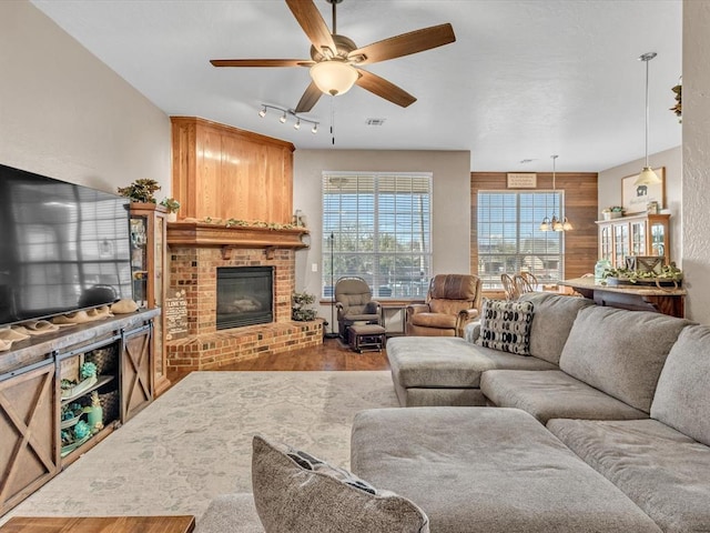 living room with wooden walls, ceiling fan with notable chandelier, a brick fireplace, and rail lighting