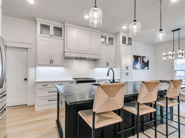 kitchen with hanging light fixtures, light wood-type flooring, a center island with sink, dark stone counters, and white cabinets