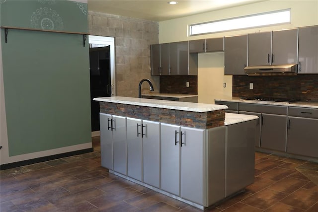 kitchen featuring decorative backsplash, under cabinet range hood, an island with sink, gray cabinetry, and gas stovetop