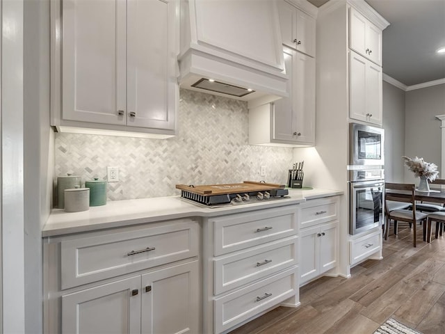 kitchen with appliances with stainless steel finishes, white cabinetry, backsplash, light hardwood / wood-style floors, and crown molding