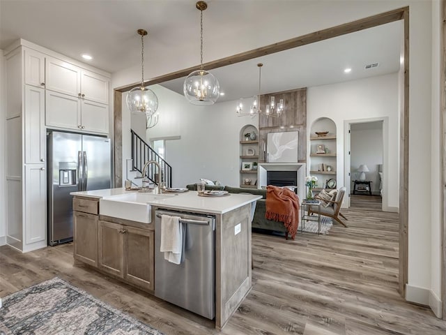 kitchen featuring appliances with stainless steel finishes, an island with sink, white cabinets, hanging light fixtures, and light wood-type flooring