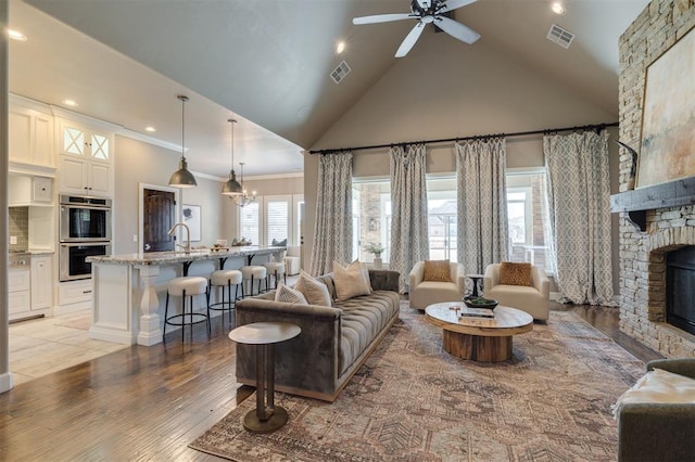 living room with a stone fireplace, high vaulted ceiling, ornamental molding, light hardwood / wood-style floors, and ceiling fan with notable chandelier