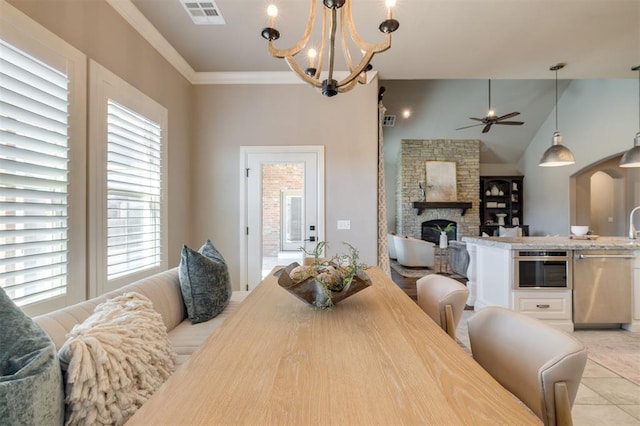 tiled dining room featuring crown molding, a stone fireplace, ceiling fan with notable chandelier, and a healthy amount of sunlight