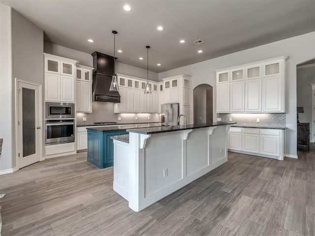 kitchen featuring decorative light fixtures, white cabinets, a large island with sink, custom exhaust hood, and stainless steel appliances