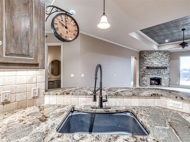 kitchen with sink, ceiling fan, a tray ceiling, ornamental molding, and a stone fireplace