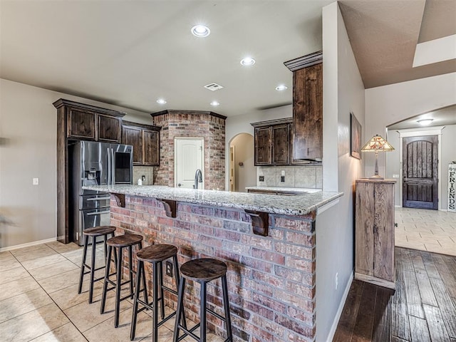 kitchen with a breakfast bar area, stainless steel fridge, backsplash, light stone counters, and dark brown cabinets