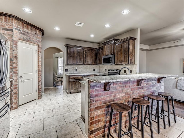 kitchen featuring a breakfast bar, appliances with stainless steel finishes, dark brown cabinets, light stone counters, and decorative backsplash