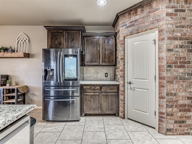 kitchen featuring stainless steel fridge, dark brown cabinetry, light stone counters, brick wall, and light tile patterned flooring