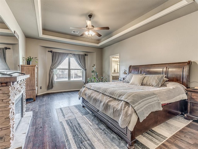 bedroom with ensuite bathroom, a stone fireplace, dark hardwood / wood-style flooring, ceiling fan, and a tray ceiling