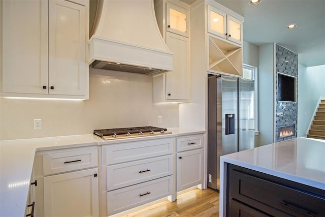 kitchen with premium range hood, appliances with stainless steel finishes, a fireplace, white cabinets, and light hardwood / wood-style floors