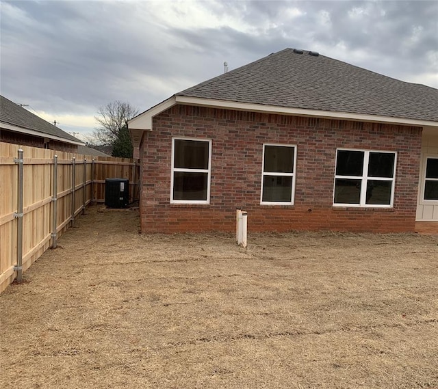 rear view of house featuring roof with shingles, fence, central AC, and brick siding