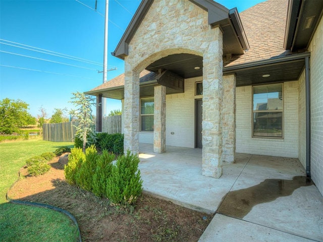 doorway to property with a patio, fence, roof with shingles, a yard, and brick siding