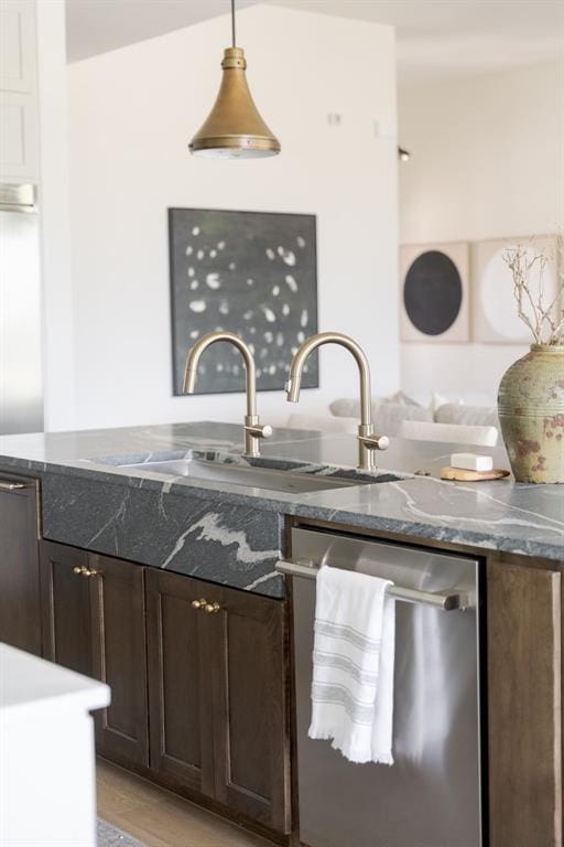kitchen featuring stainless steel dishwasher, a sink, dark stone counters, dark brown cabinetry, and decorative light fixtures