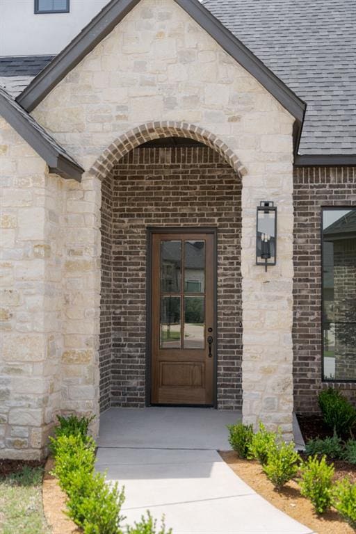 doorway to property featuring brick siding, stone siding, and a shingled roof