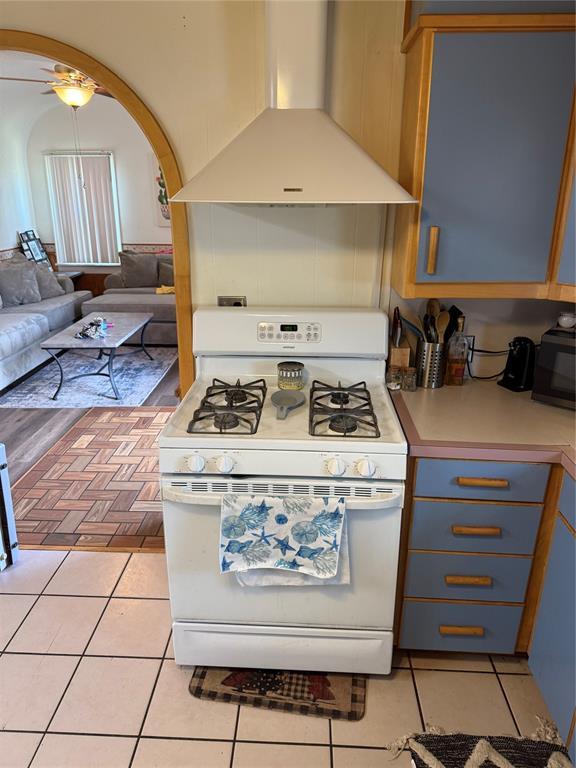 kitchen with range hood, white gas stove, blue cabinetry, light tile patterned floors, and light countertops