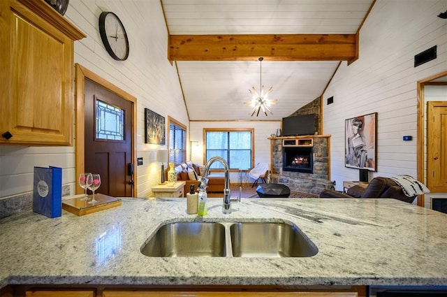 kitchen featuring sink, vaulted ceiling with beams, a notable chandelier, a fireplace, and light stone countertops