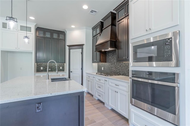 kitchen featuring premium range hood, sink, appliances with stainless steel finishes, a kitchen island with sink, and white cabinets