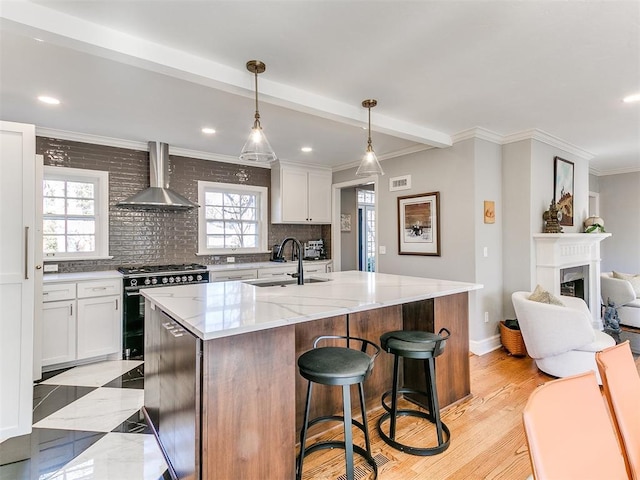 kitchen with sink, white cabinetry, an island with sink, black gas range, and wall chimney range hood