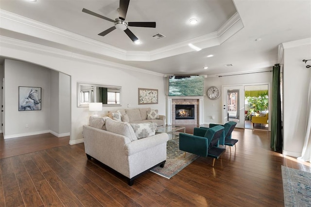 living room with dark wood-type flooring, crown molding, a tray ceiling, ceiling fan, and a fireplace