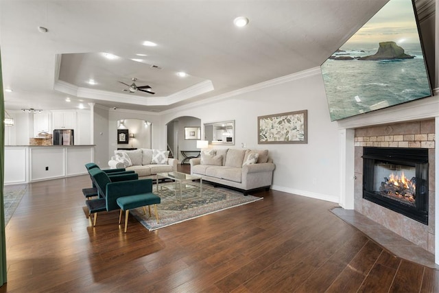 living room featuring dark wood-type flooring, ceiling fan, a tray ceiling, ornamental molding, and a tiled fireplace