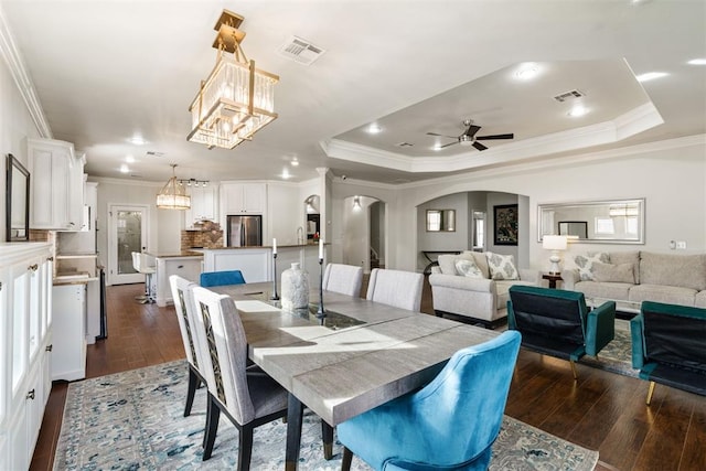 dining room with ornamental molding, dark wood-type flooring, ceiling fan with notable chandelier, and a tray ceiling