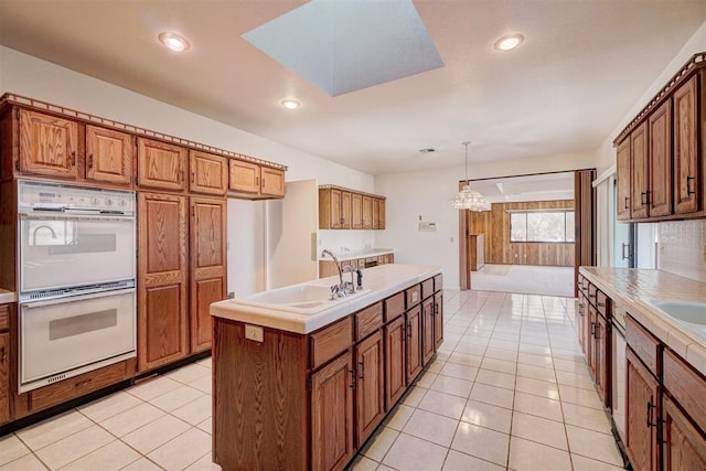 kitchen with sink, light tile patterned floors, white double oven, a center island with sink, and decorative light fixtures