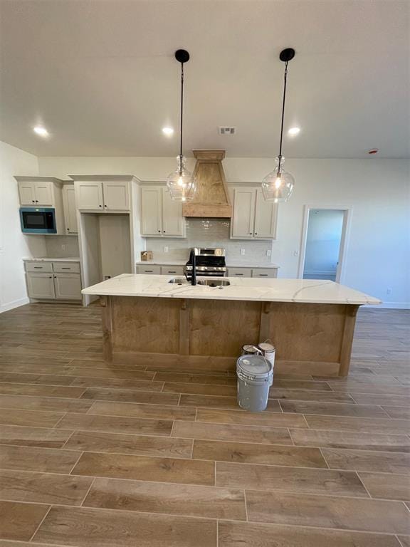 kitchen with sink, custom exhaust hood, hanging light fixtures, a large island with sink, and light stone counters