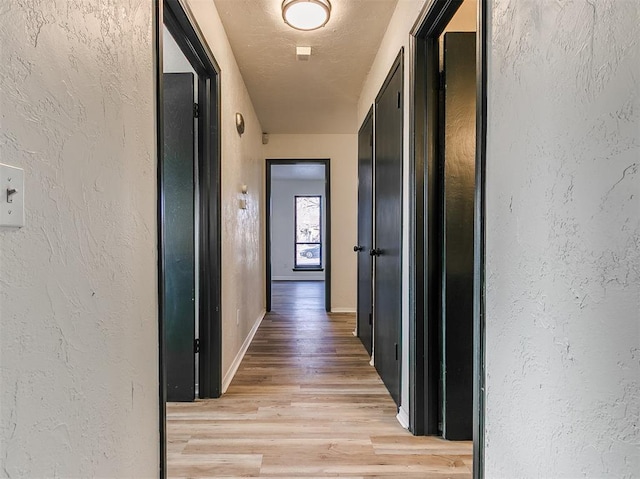 corridor with a textured ceiling, a textured wall, light wood-type flooring, and baseboards