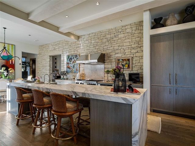 kitchen with dark wood-type flooring, wall chimney exhaust hood, beam ceiling, decorative light fixtures, and a large island