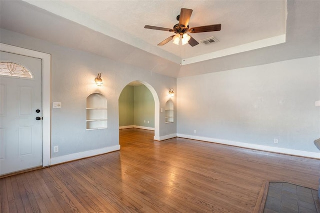 empty room with a raised ceiling, wood-type flooring, ceiling fan, and built in shelves