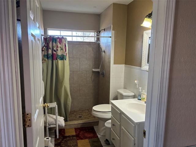 bathroom with toilet, vanity, tile walls, tiled shower, and wainscoting