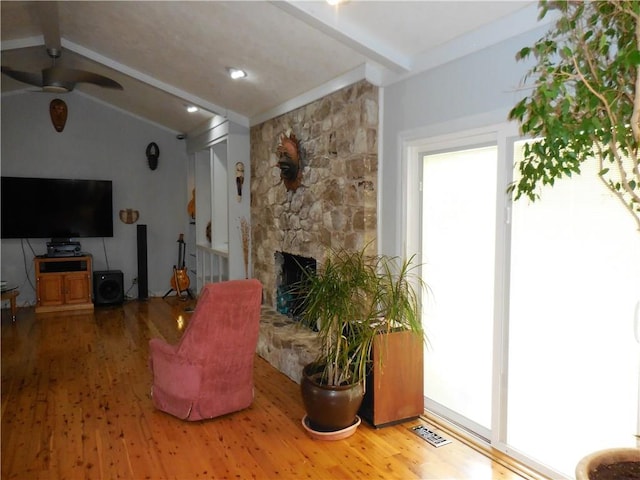 living room featuring hardwood / wood-style flooring, ceiling fan, a stone fireplace, and lofted ceiling with beams