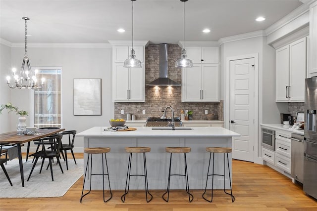 kitchen featuring wall chimney range hood, sink, a kitchen island with sink, white cabinetry, and hanging light fixtures