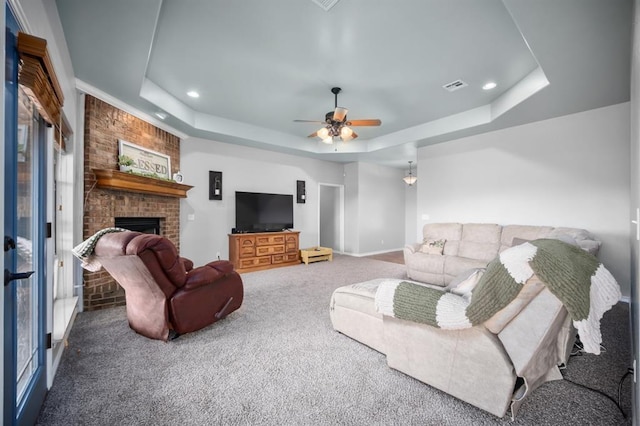 carpeted living room with a brick fireplace, ceiling fan, and a tray ceiling