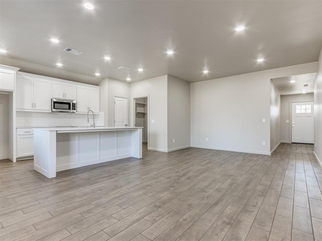 kitchen featuring a kitchen island with sink, white cabinets, backsplash, and light wood-type flooring