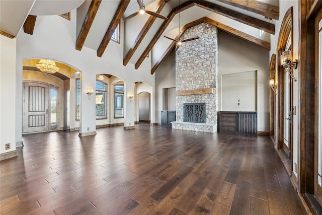 unfurnished living room featuring ceiling fan with notable chandelier, beam ceiling, high vaulted ceiling, dark hardwood / wood-style floors, and a stone fireplace